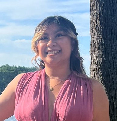 photo of Aimee Layo, Filipino woman with brown hair in a light reddish pink jumpsuit, standing outside in front of a body of water leaning against a wooden fence