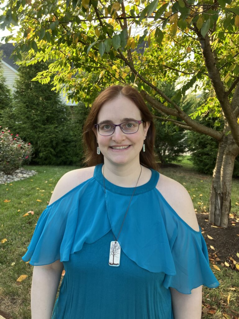 photo of Dr. Sarah Norton, white woman with brownish reddish hair wearing a light blue top standing outside on grass in front of a lush green tree