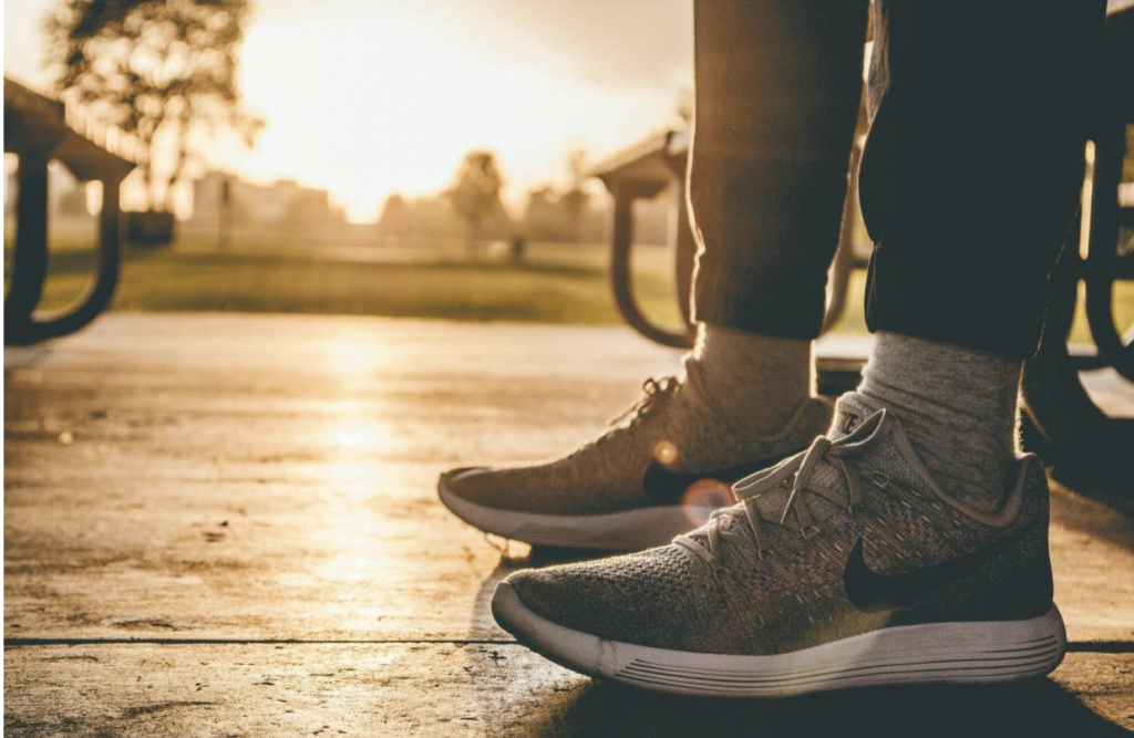 a photo of someone standing outside from the knee down, wearing gray running shoes, gray socks, and black athletic pants, lit by golden sunlight that looks like sunrise.