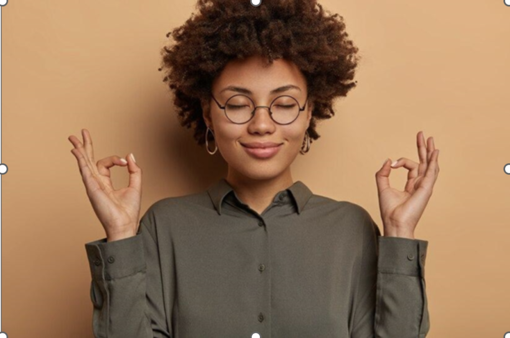 a smiling black woman with an afro and glasses in an earthy dark brown long-sleeve shirt stands holding both hands in the gyan mudra against a tan background