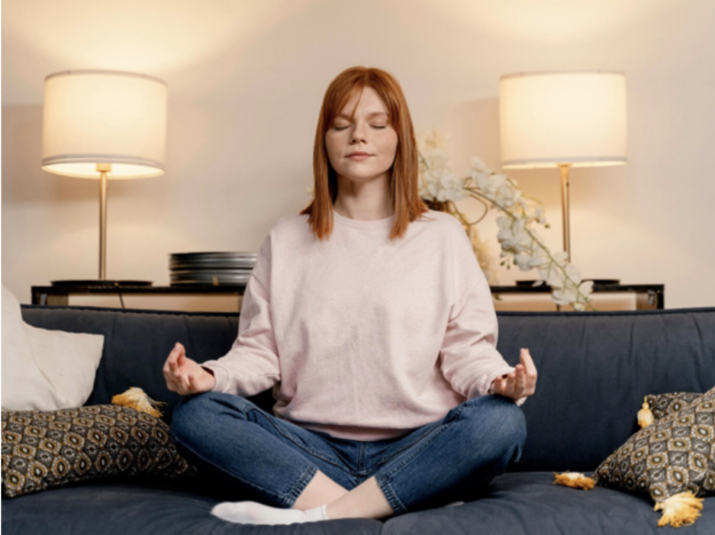 red headed white woman wearing a pink sweatshirt and jeans sits in a cross-legged meditation position on a couch. She has her eyes closed, a slight smile, and her fingers hands resting on her knees in the mudra position.