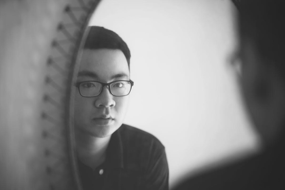 Asian man with black-rimmed glasses looks at his reflection in the mirror.