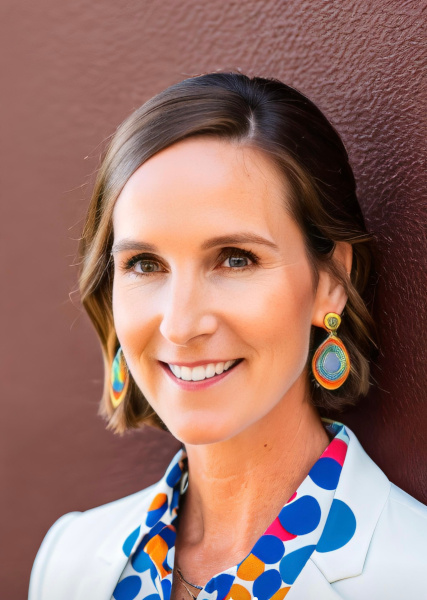 white woman with brown hair and white shirt with colorful blue and pink collar and colorful dangling earrings smiles