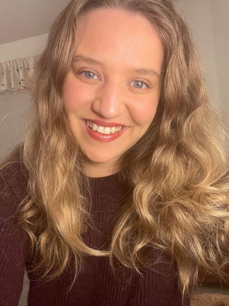 a woman in a maroon sweater with dirty blonde hair, blue eyes, and light red lipstick smiles wide, teeth exposed.